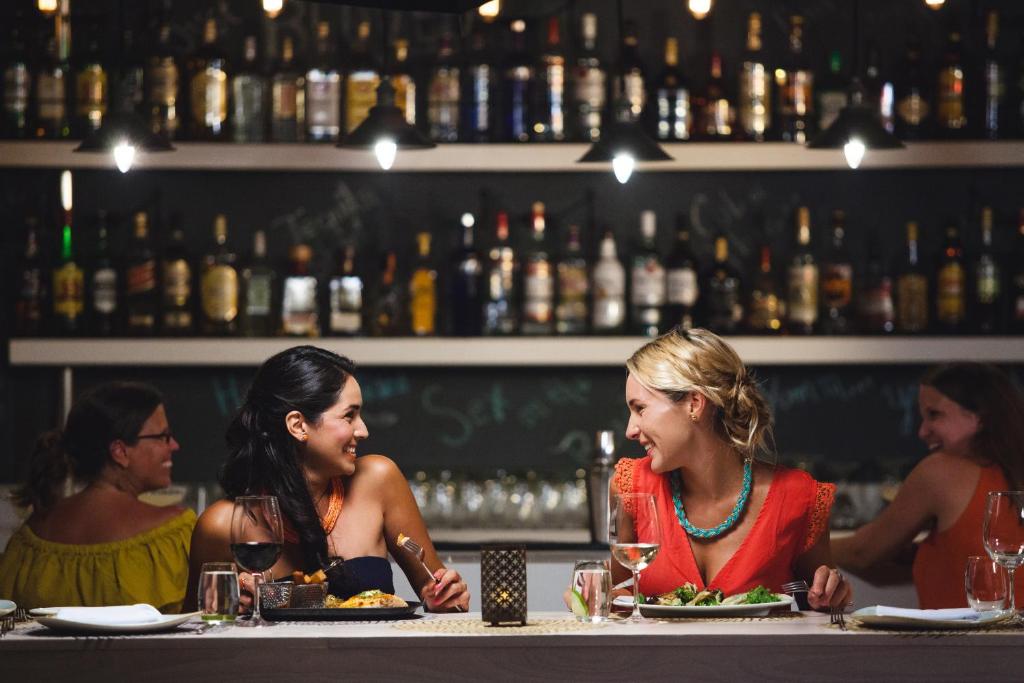 Divas sievietes sēž pie galda restorānā, baudot vakariņas un sarunājoties, kamēr fona plauktā redzami dažādi alkoholiskie dzērieni. Gaisma ir silta un atmosfēra ir draudzīga.