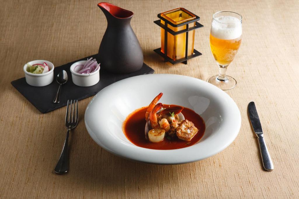 Gastronomiskais ēdiens ar garnelēm un kalmāriem, pasniegts elegantā baltā traukā, blakus glāze alus un dekoratīva lampiņa.