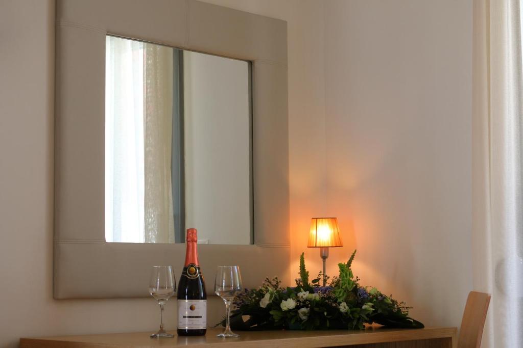 Šampanieša pudele un glāzes uz koka galda, blakus ziedu kompozīcija un maiga lampiņa, pie sienas liels spogulis.