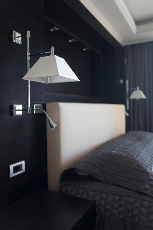 Mūsdienīga guļamistaba ar elegantu gultas galvgali un stilīgām lampām, kas piekārtas pie tumša sienas. Interjers ir minimālistisks un ērts, radot relaksējošu atmosfēru.