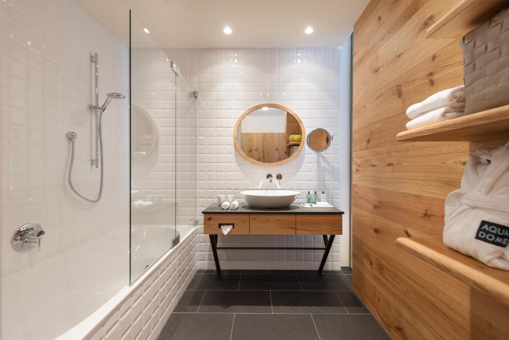 Moderns vannas istabas interjers ar stikla dušu, baltiem flīzēm un koka akcentiem, kas piešķir siltumu un eleganci. Vannas istabā ir mūsdienīga izlietne ar apaļu spoguli un plauktiņš ar dvieļiem un higiēnas līdzekļiem.
