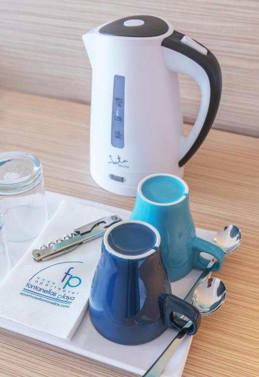 Tējkanna un krūzes uz galda, kopā ar karotēm un salveti, veido mājīgu tējas stūrīti. Baltā tējkanna ar melnu rokturi un divas krāsainas krūzes papildina koka virsmu.