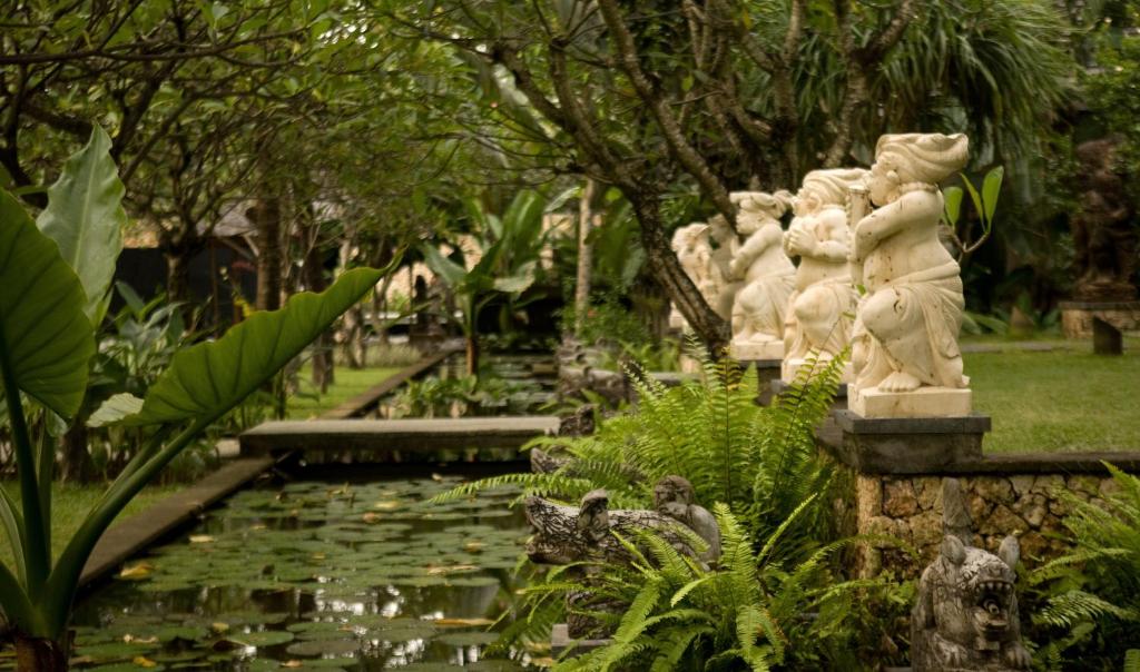 Dārzs ar zaļām lapām un skaistām skulptūrām, kas izkārtotas gar dīķi. Attēlā redzami arī tropiskie augi un ūdens lilijas.