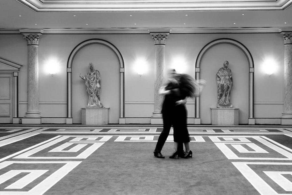 Skaista melnbalta fotogrāfija, kurā redzami divi dejotāji elegantā telpā ar greznām skulptūrām un izsmalcinātu grīdas dizainu.