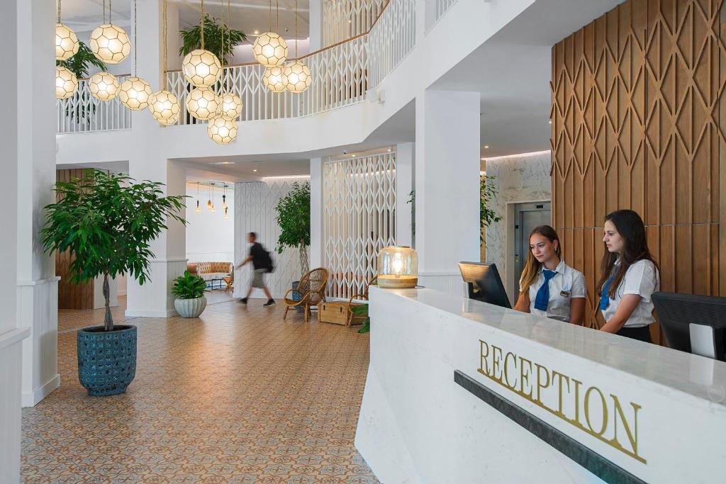 Modernas viesnīcas reģistratūra ar stilīgu interjeru, kas ietver augu dekorus un elegantu apgaismojumu. Divas viesnīcas darbinieces apkalpo viesus pie reģistratūras.