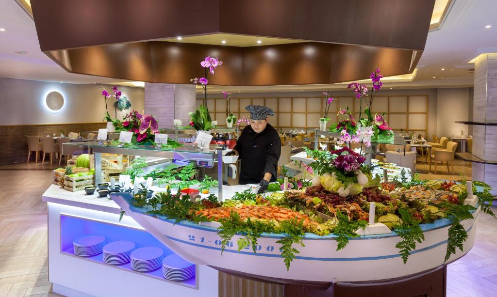 Elegants restorāns ar krāsainu bufeti, kurā redzams šefpavārs, kas strādā pie jūras velšu un dārzeņu galda, ko rotā ziedi un zaļumi. Iekārtojums ir moderns un viesmīlīgs.