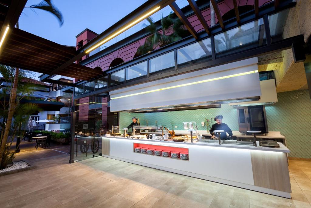 Mūsdienīgs atvērts restorāns ar šefpavāru, kas gatavo ēdienus pie moderna galda. Apkārt ir zaļumi un terase, radot mājīgu atmosfēru.