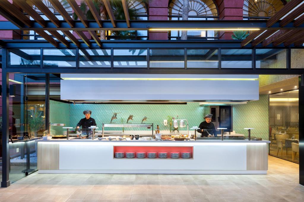 Mūsdienīga virtuve ar šefpavāriem, kas strādā pie atvērtā bufetes stila galda. Interjers ir gaišs un stilīgs, ar uzsvaru uz skaidrām līnijām un moderniem elementiem.