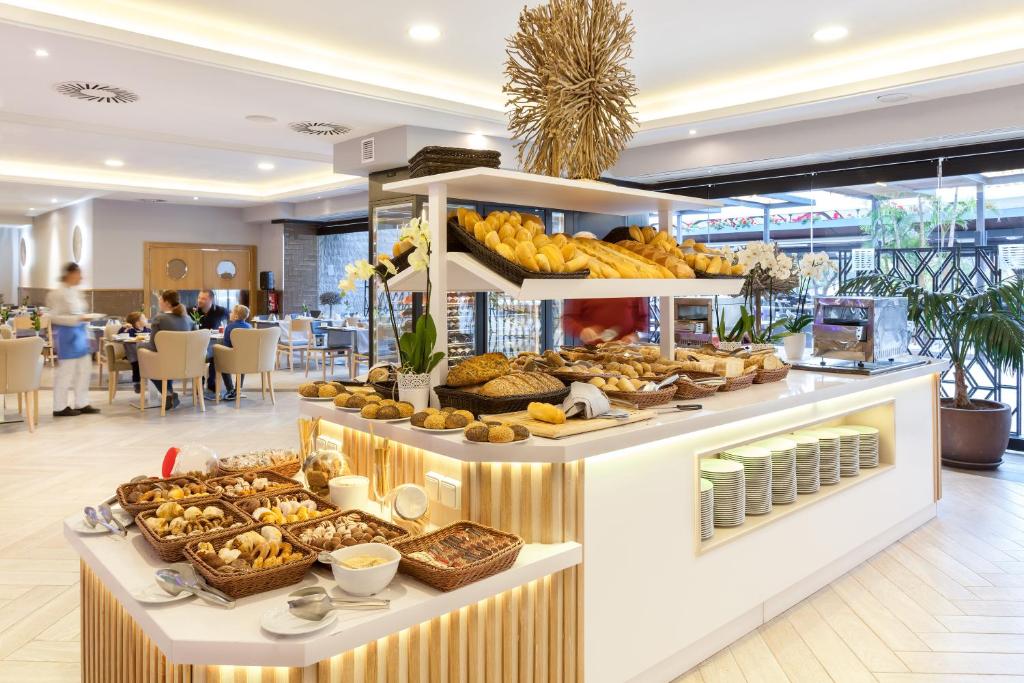 Brokastu bufete ar dažādiem maizes izstrādājumiem un konditorejas izstrādājumiem, apgaismota mūsdienīgā restorānā ar elegantu interjeru. Klientu sēdvietas ir redzamas fonā, radot patīkamu atmosfēru.