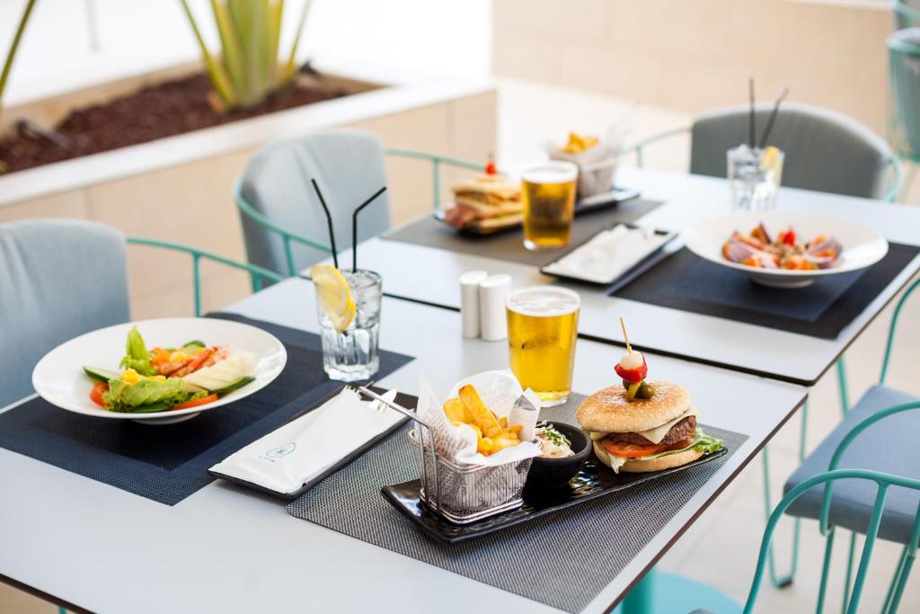 Svaigākie ēdieni un dzērieni uz galda, tostarp burgeris ar frī kartupeļiem, salāti un auksts alus. Moderns restorāna interjers ar gaišām krāsām un patīkamu atmosfēru.