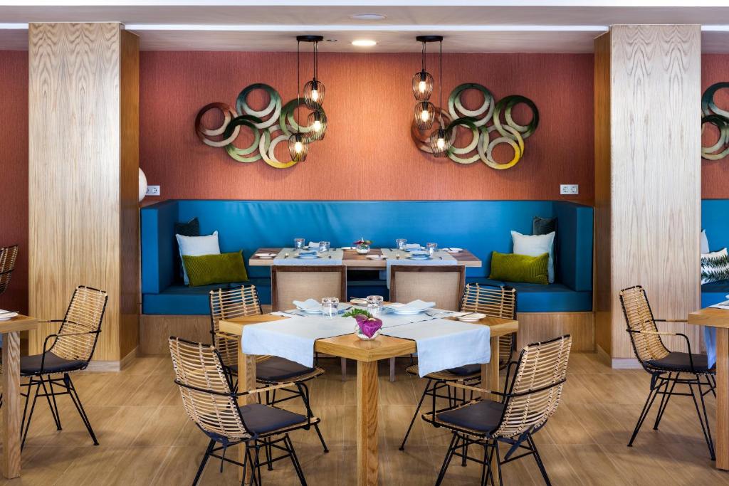 Mūsdienīgs restorāna interjers ar koka mēbelēm, zilu sēdvietu un dekoratīviem gaismas ķermeņiem, radot siltu un aicinošu atmosfēru. Galdi ir sakārtoti ar elegantiem galda piederumiem.