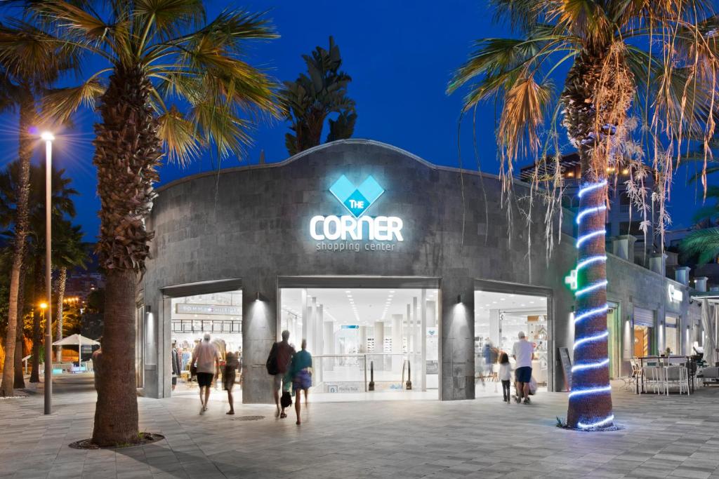 Mūsdienīga iepirkšanās centra fasāde ar palmām un izgaismotiem elementiem, kas izceļ atmosfēru vakara stundās.