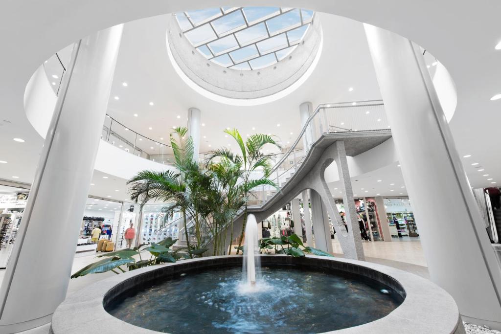 Mūsdienīgs iepirkšanās centrs ar ūdens strūklaku un zaļajiem augiem centrā, kas papildina telpas estētiku un gaisīgumu. Gaisma ieplūst no jumta loga, radot patīkamu atmosfēru.