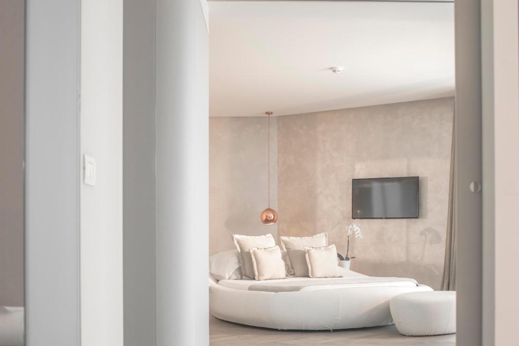 Mūsdienīgs guļamistabas interjers ar apaļu gultu, spilveniem un televizoru uz sienas. Telpa ir gaiša un eleganta, ar minimālistisku dizainu.