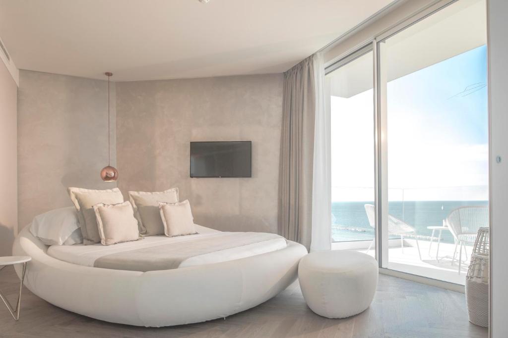 Mūsdienīgs guļamistabas interjers ar apaļu gultu, elegantām spilveniem un skatu uz jūru caur lieliem logiem. Telpa ir gaiša un gaumīgi iekārtota, radot relaksējošu atmosfēru.