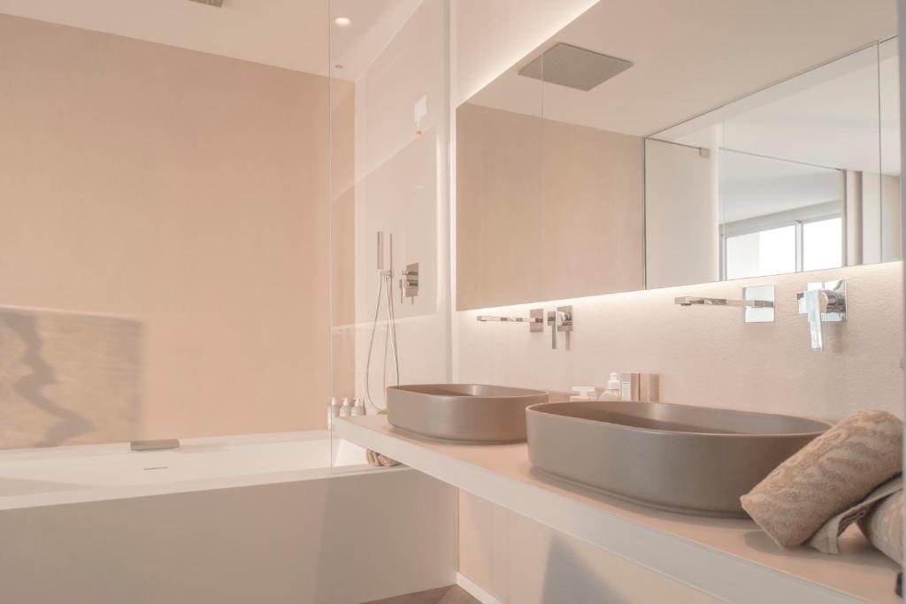 Mūsdienīgs vannas istabas interjers ar divām pelēkām izlietnēm un elegantu spoguli, kas atspoguļo gaismas un telpas plašumu. Vannas istaba ir dekorēta ar neitrālām krāsām un minimālistisku dizainu.