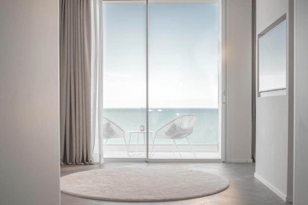 Mūsdienīgs interjers ar skatu uz jūru, kurā redzams apaļš paklājs un divas krēsli terasei. Gaisma plūst cauri lielajiem logiem, radot mierīgu atmosfēru.