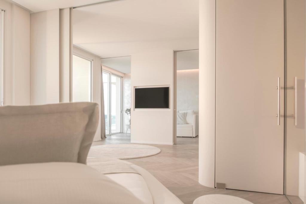 Mūsdienīga guļamistabas interjera skats ar gaišām krāsām un atvērtu plānojumu, kur redzams televizors un piekļuve citai telpai.