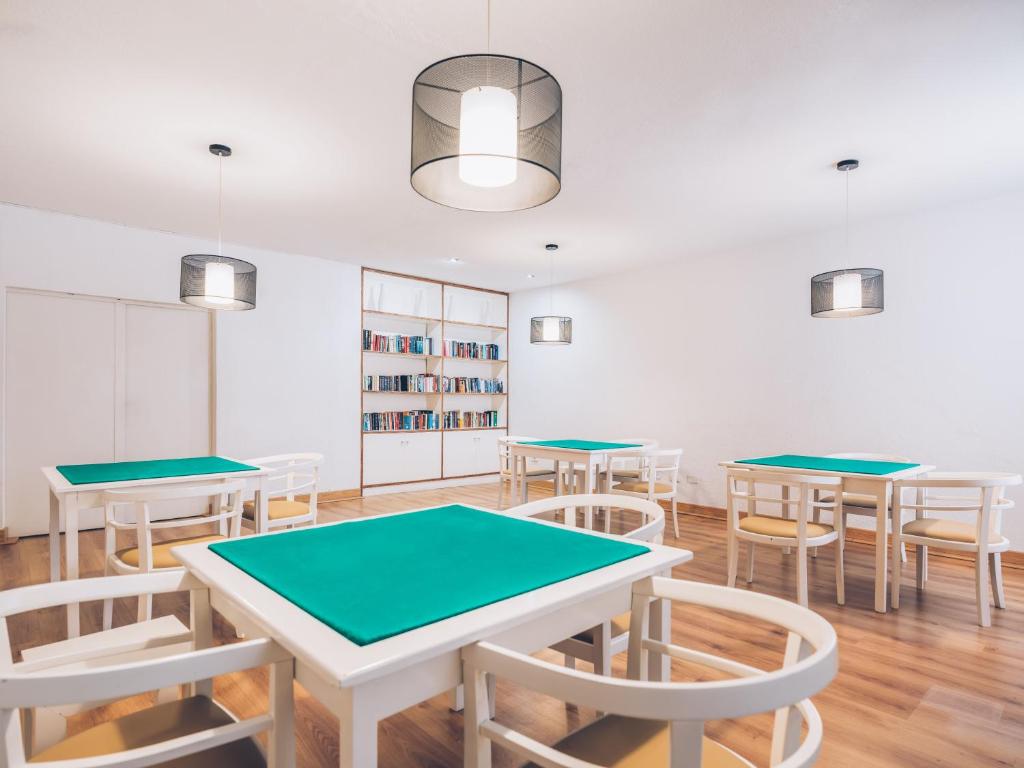 Gaiša un moderna telpa ar baltām galdiem un zaļu virsmu, kurā ir vairākas sēdvietas. Uz sienas atrodas grāmatu plaukts, kas piešķir telpai mājīgu atmosfēru.