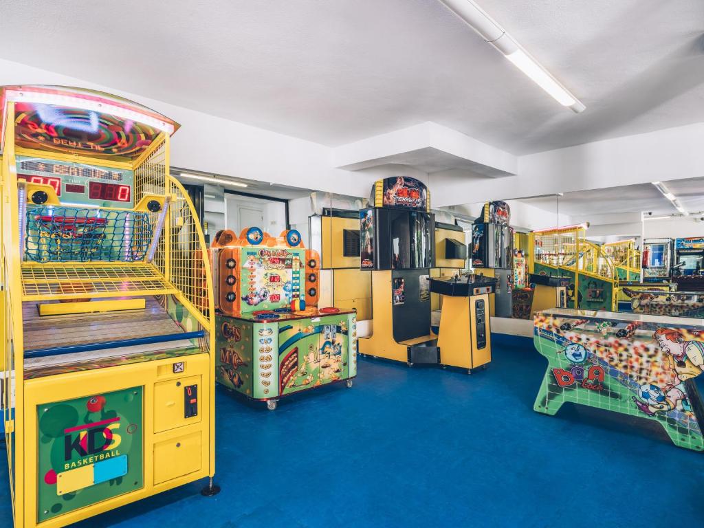 Arcade spēļu telpa ar dažādām spēļu automātiem, tostarp basketbola un citu interaktīvo spēļu iekārtām. Dzīvespriecīga atmosfēra un krāsaini elementi.