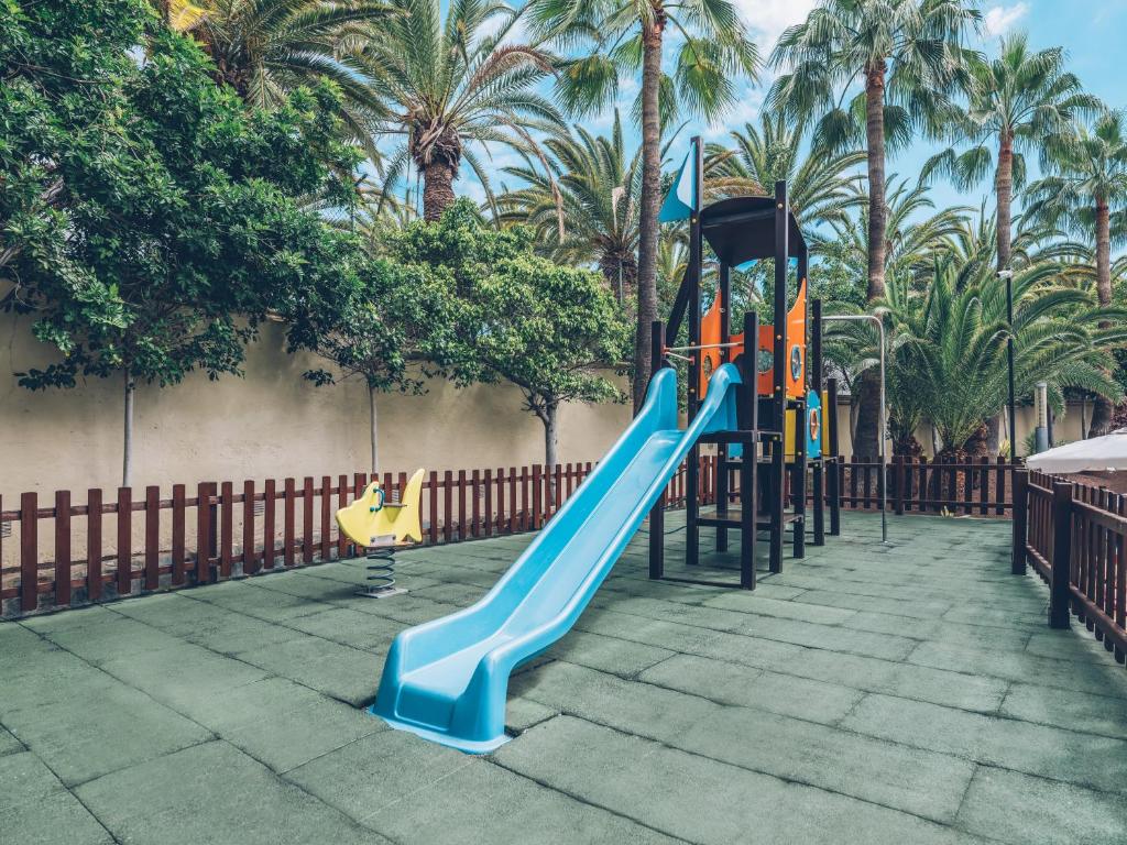 Bērnu rotaļu laukums ar zilu slidkalniņu un dzeltenu šūpuli, apkārt palmām un zaļām lapām. Ideāla vieta bērniem spēlēties un baudīt dabu.