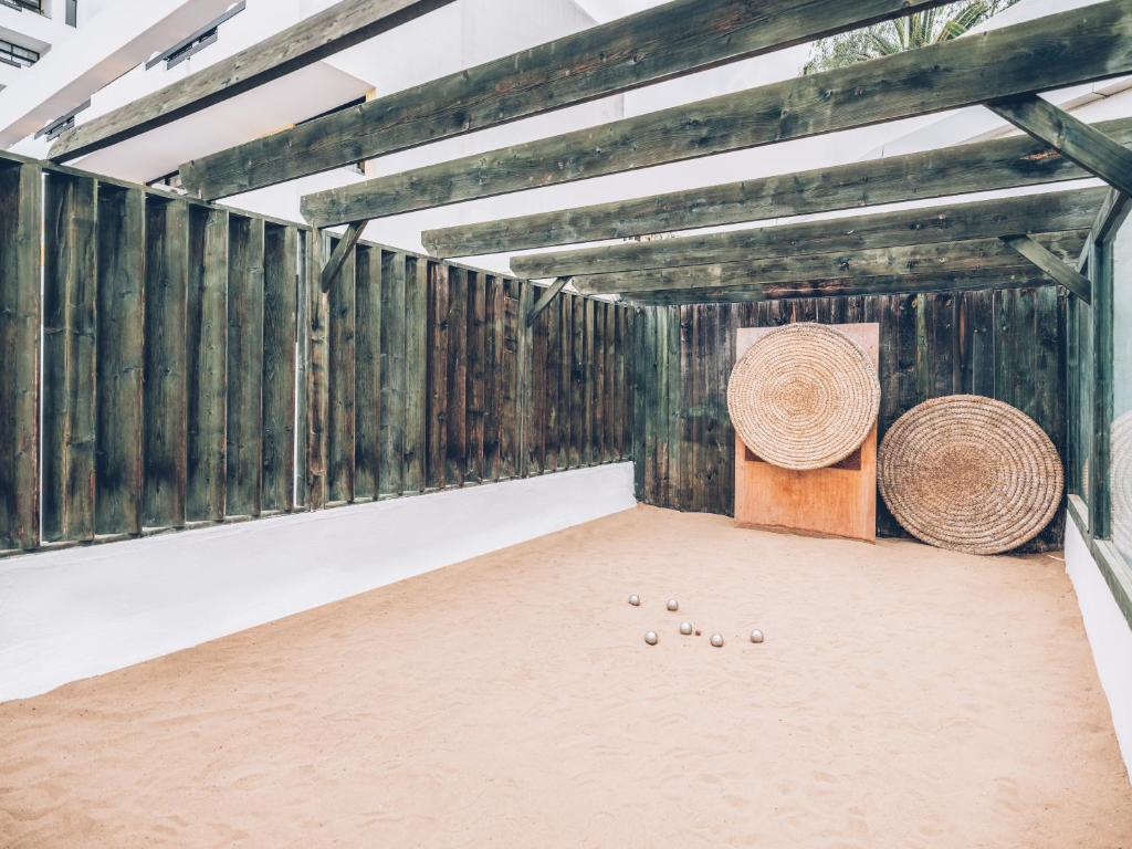 Sakņu laukums ar smiltīm un koka konstrukcijām, kurā redzami bumbas un divi lieli, apaļi, aušanas elementi. Ideāli piemērots boules spēlei vai relaksācijai.