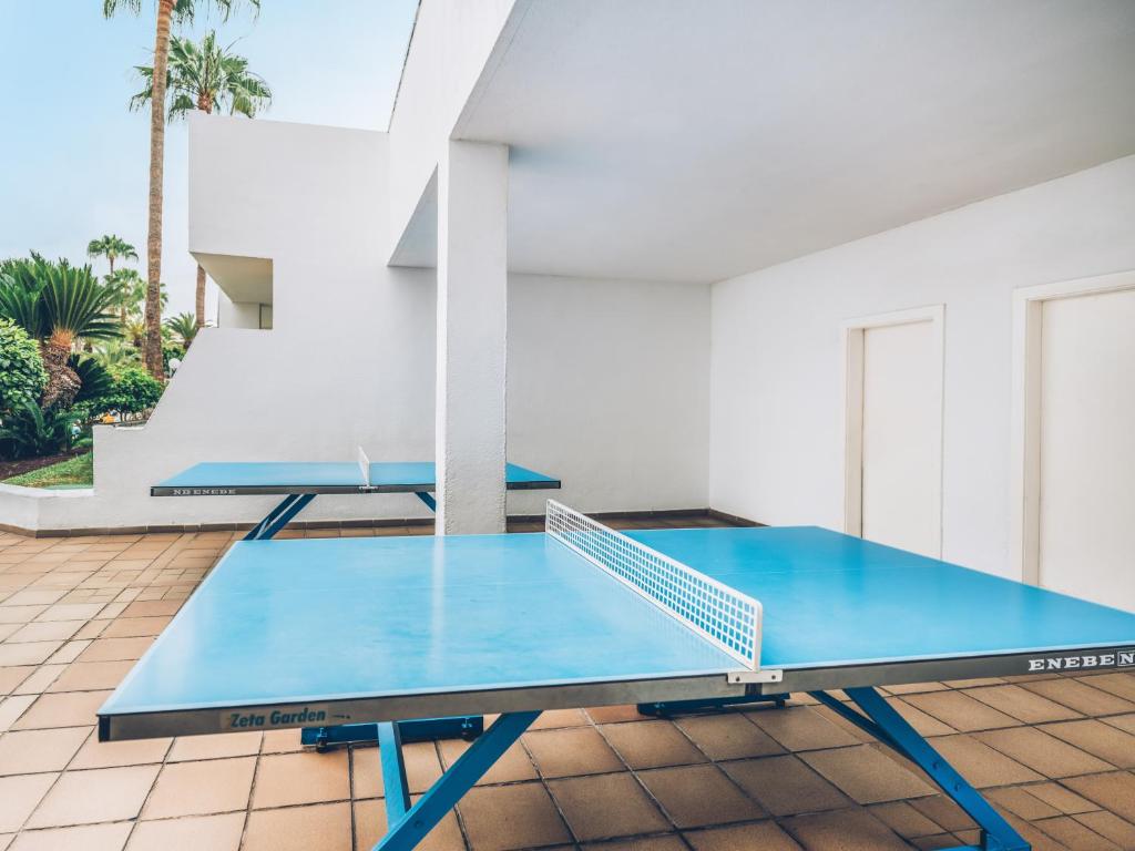 Pingponga galdi sporta zona ar zilu virsmu un tīklu, novietota ārpusē, apkārt zaļām augiem un baltiem sienām.