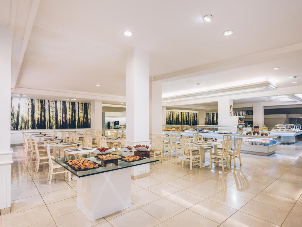 Modernas restorāna iekārtojums ar gaišām krāsām un koka mēbelēm, plašs ēdināšanas laukums ar bufeti un dekoratīvām ainavām uz sienām.