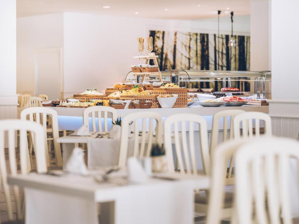 Mūsdienīgs restorāns ar plašu bufeti, kas piedāvā dažādus ēdienus un uzkodas, ar baltām mēbelēm un patīkamu atmosfēru.