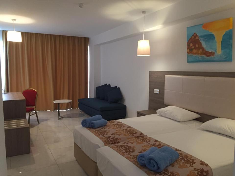 Mūsdienīgs viesnīcas numurs ar divām gultām, dīvānu un rakstāmpalīgu. Interjeru papildina gaismas un mākslas darbs uz sienas.