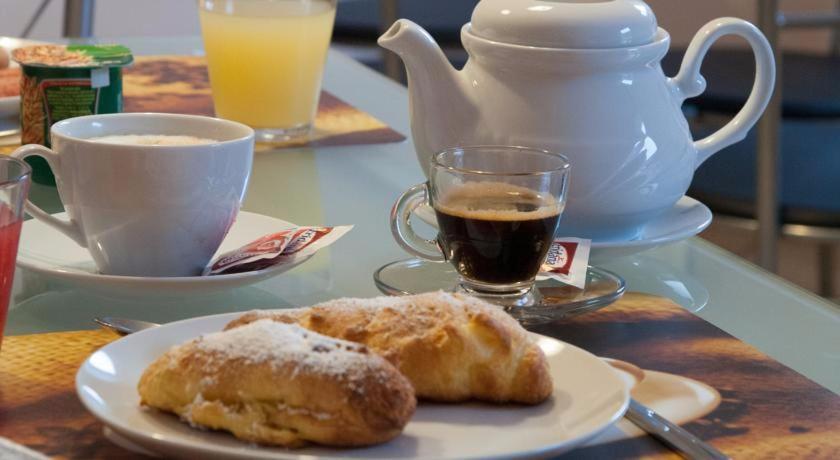 Brokašu galda iestatījums ar keramikas tējkannu, kafijas krūzi un garšīgiem cepumiem, kas pārkaisīti ar cukura pulveri. Fons sastāv no dzērieniem, tostarp apelsīnu sulas un kafijas.