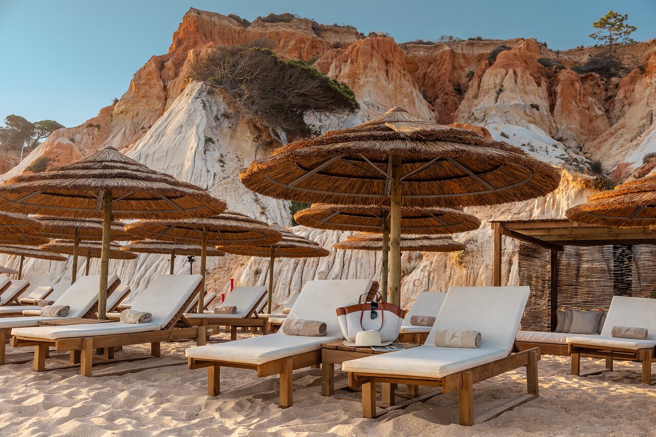 EPIC SANA Algarve Hotel pludmales zona ar ērtiem koka sauļošanās krēsliem un salmu saulessargiem pie sarkanajām klintīm.