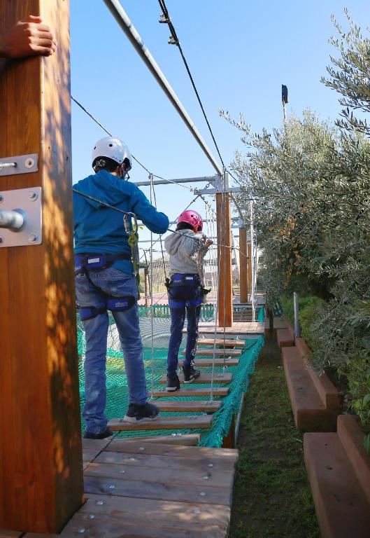 Divi bērni izpēta piedzīvojumu parku, ejot pa virvju tiltu, tērpti drošības aprīkojumā un ķiverēs. Fons ir skaidrs debesis un zaļo krūmu rinda.
