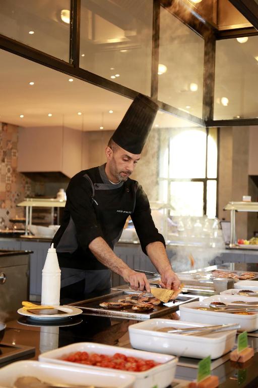 Šefpavārs gatavo ēdienus stilīgā virtuvē, pievēršoties grilēšanai un radošai prezentācijai. Apkārt ir dažādi ēdieni un garšvielu konteineri.