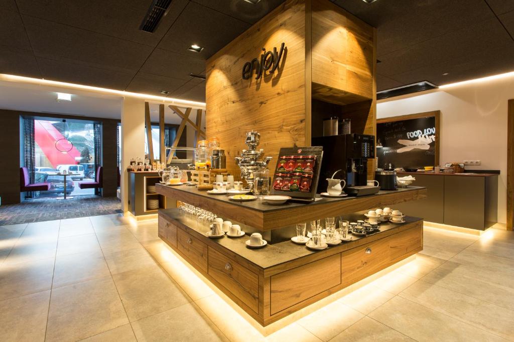 Stilīgs kafejnīcas stūris ar koka akcentiem, kurā redzami dažādi dzērieni un kafijas automāts, apgaismots ar siltu gaismu. Interjers ir moderns un aicinošs.