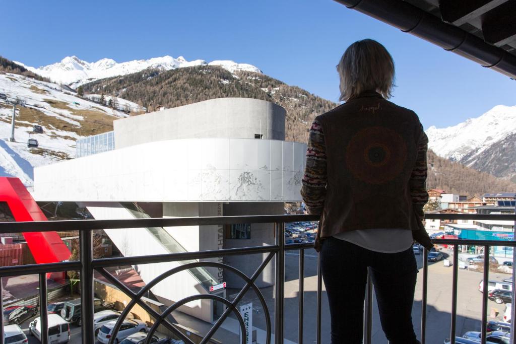 Sieviete ar īsām matu frizūrām stāv uz balkona, skatoties uz sniegotajiem kalniem un modernu ēku. Fons iezīmē ziemas ainavu ar slēpošanas pacēlājiem un pilsētas dzīvi.