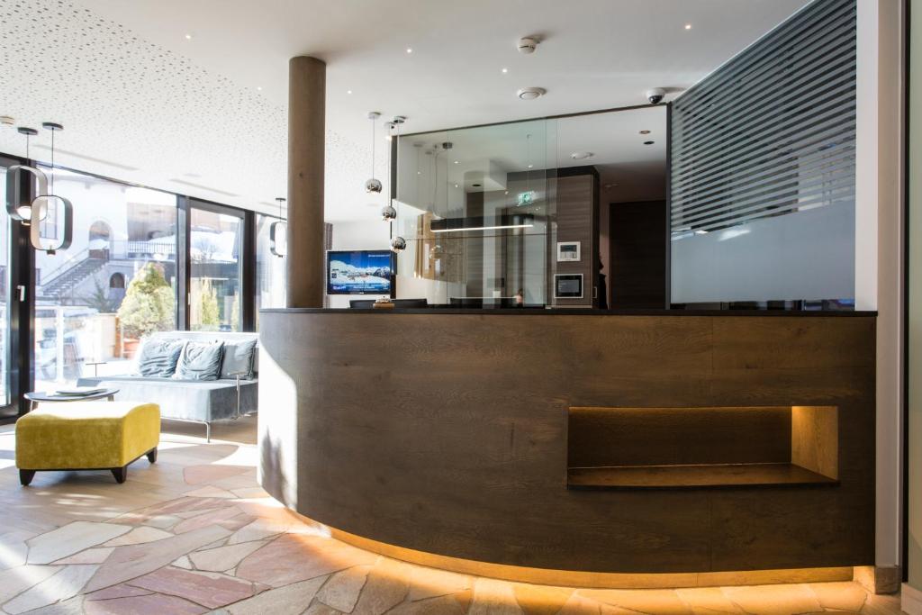 Mūsdienīgs viesnīcas reģistratūras interjers ar koka un stikla elementiem, kurā redzama dīvāna zona un gaiss, kas ieplūst caur lieliem logiem.