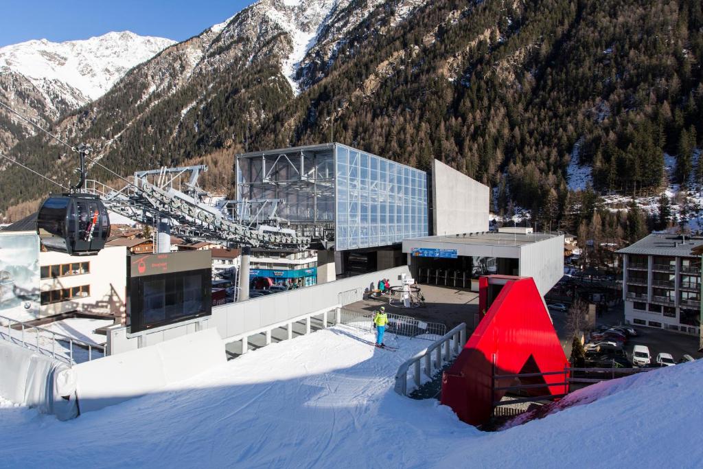 Mūsdienīga kalnu slēpošanas pacēlāja stacija ar modernu arhitektūru un sniegotu ainavu, kas redzama kalnu fonā. Sniega seguma un kalnu apkārtne rada ideālu vidi ziemas sporta aktivitātēm.