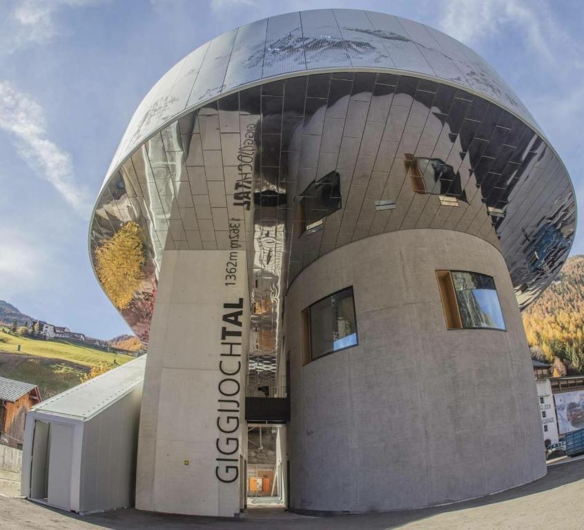 Mūsdienīga ēka ar spīdīgu metāla jumtu un betona sienām, kas atrodas kalnu apvidū, ar uzrakstu "Giggijochtal" un informāciju par augstumu. Apkārtne tiek piepildīta ar zeltainiem koku lapām un kalnu ainavām.
