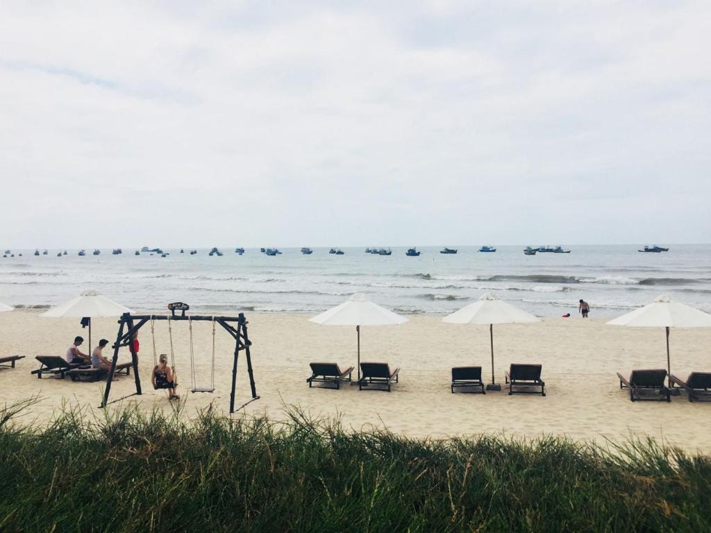 Pludmales aina ar saulessargiem, šūpolēm un zvejas laivām jūrā. Rāmā atmosfērā cilvēki atpūšas uz sauļošanās krēsliem.