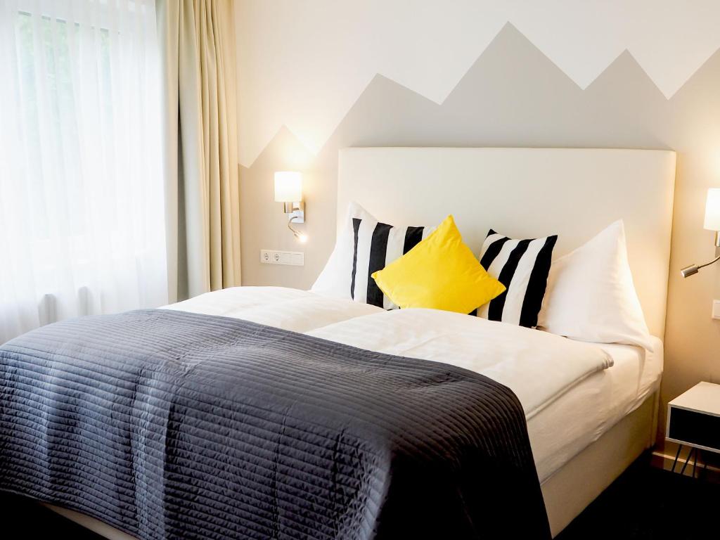 Mūsdienīgs guļamistabas interjers ar divguļamo gultu, melnu un baltu svītrainu spilvenu, dzeltenu dekoratīvo spilvenu un mīkstu, pelēku segu. Fons rotāts ar stilizētiem kalnu motīviem.