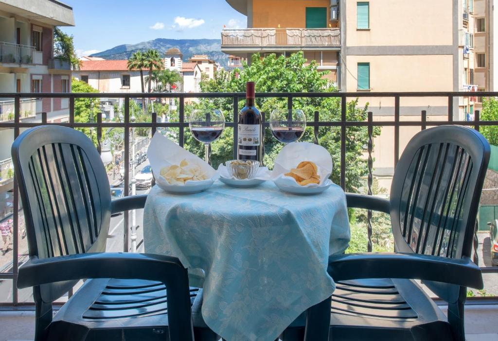 Balkona galds ar divām plastmasas krēslām, uz kura stāv vīna pudele un divi glāzes ar vīnu, kā arī čipsi. Fons ir pilsētas ainava ar kokiem un ēkām.