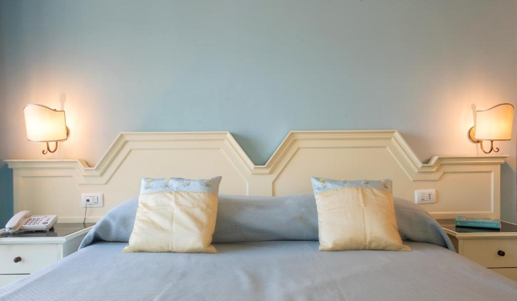 Mūsdienīgs guļamistabas interjers ar gaiši zilu sienu, elegantu gultas galvu un mīkstiem spilveniem. Sānos redzami sienas lampiņas un telefons uz naktsgaldiņa.