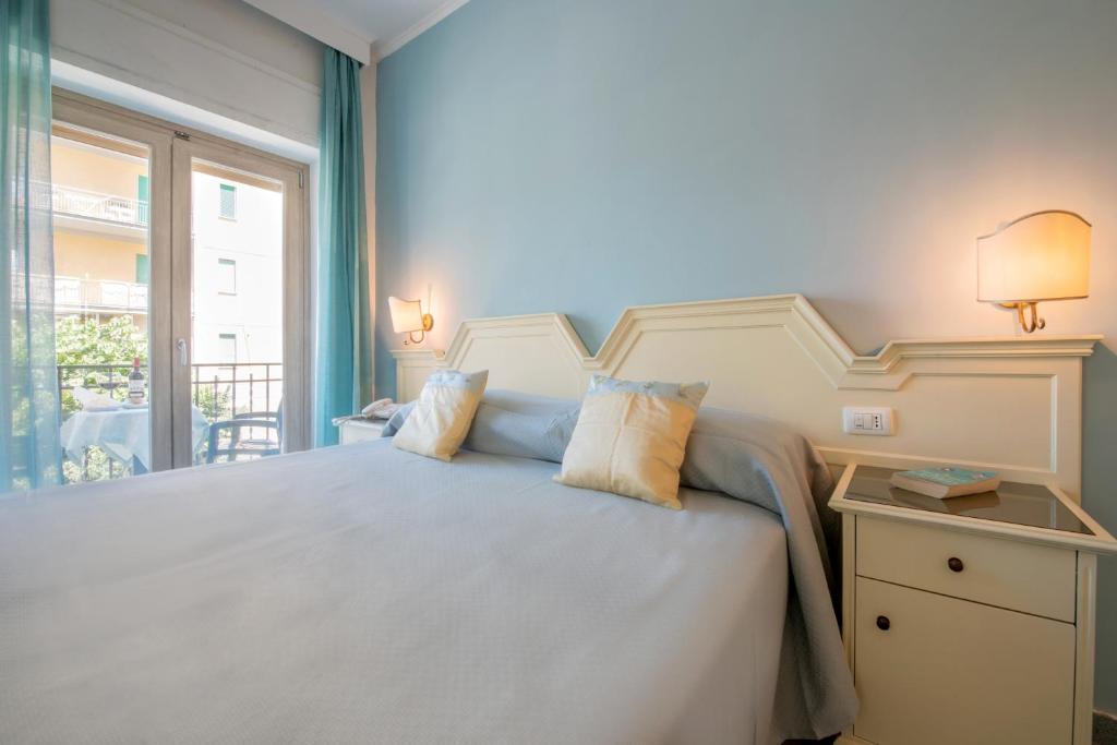 Gaiša guļamistaba ar zilu sienu, divguļamo gultu un mīkstiem spilveniem. Logi ar caurspīdīgām aizkariem ved uz balkonu, kur redzama dārza aina.