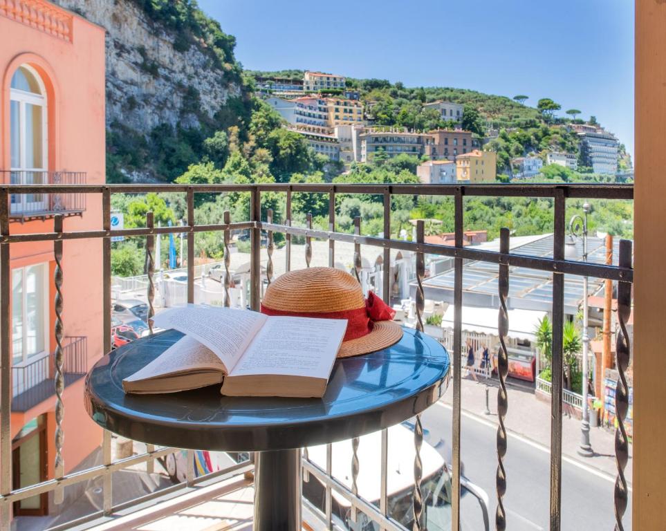 Saulaina balkona skats ar grāmatu un cepuri uz galda, apkārt redzamas kalnu ainavas un pilsētas dzīve. Ideāla vieta relaksācijai un lasīšanai.