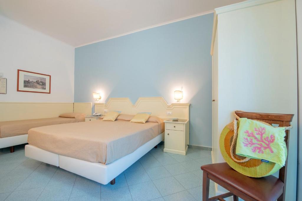 Gaišā viesnīcas numuriņa iekārtojums ar divām gultām un gaišām sienām, kas rada mājīgu atmosfēru. Uz krēsla atpūšas krāsaina rokas darbs.