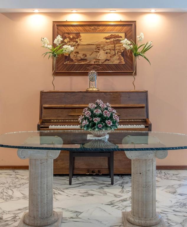 Interjera elements ar klavierēm, stikla galdu un puķu kompozīciju, kas papildināts ar gleznu uz sienas.