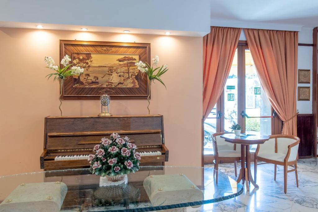 Eleganta viesnīcas uzgaidāmā telpa ar klavierēm un mākslas darbu pie sienas, dekorēta ar ziediem un mīkstiem aizkariem. Sēžamā zona ar galdu un krēsliem rada mājīgu atmosfēru.