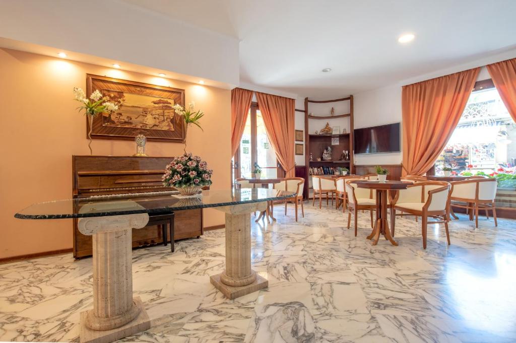 Eleganta viesistaba ar marmora grīdu, stikla galdu un tradicionālu klavieri, apgaismota ar siltu gaismu un dekorēta ar ziediem. Logi ar oranžiem aizkariem paver skatu uz ārpusi.