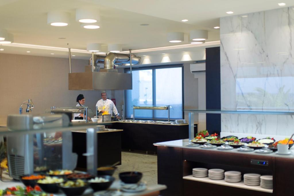 Mūsdienīga virtuve ar šefpavāriem, kas gatavo ēdienus, un dažādām uzkodām uz galda. Interjers ir gaišs un elegants, radot patīkamu atmosfēru.